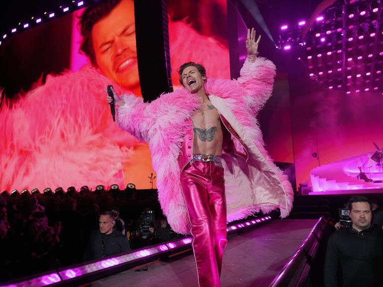 Harry Styles performt auf der Bühne. Er trägt einen Mantel aus pinkfarbenen Federn.