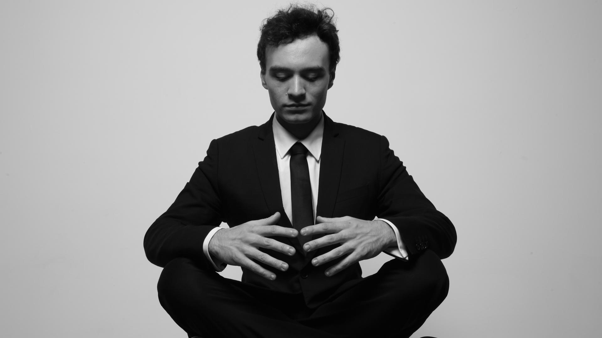 Ein junger Mann sitzt im Schneidersitz und blickt konzentriert auf seine Hände. Er trägt einen schwarzen Anzug. Es ist der französische Pianist Jonathan Fournel.