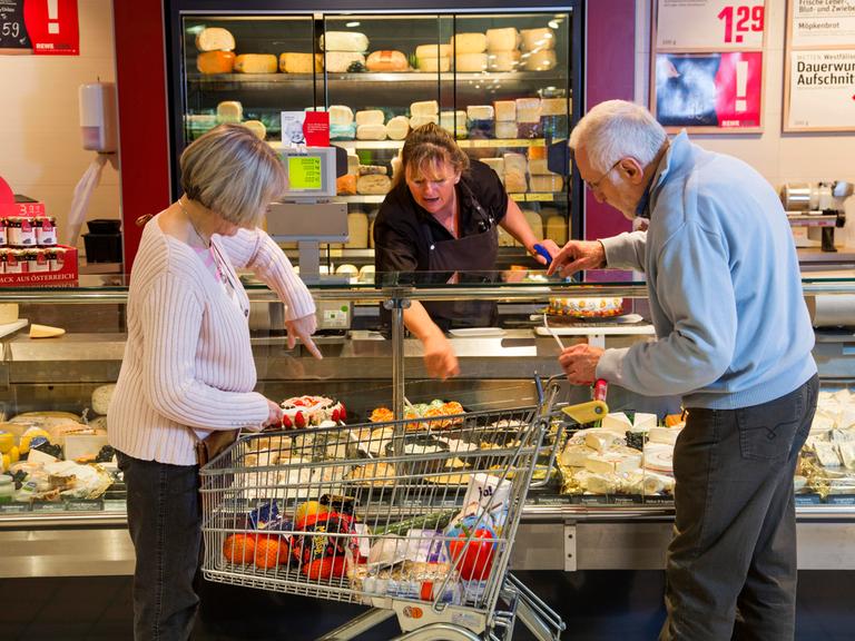 Seniorenpaar mit Einkaufswagen beim Einkaufen im Supermarkt, an der Käsetheke.