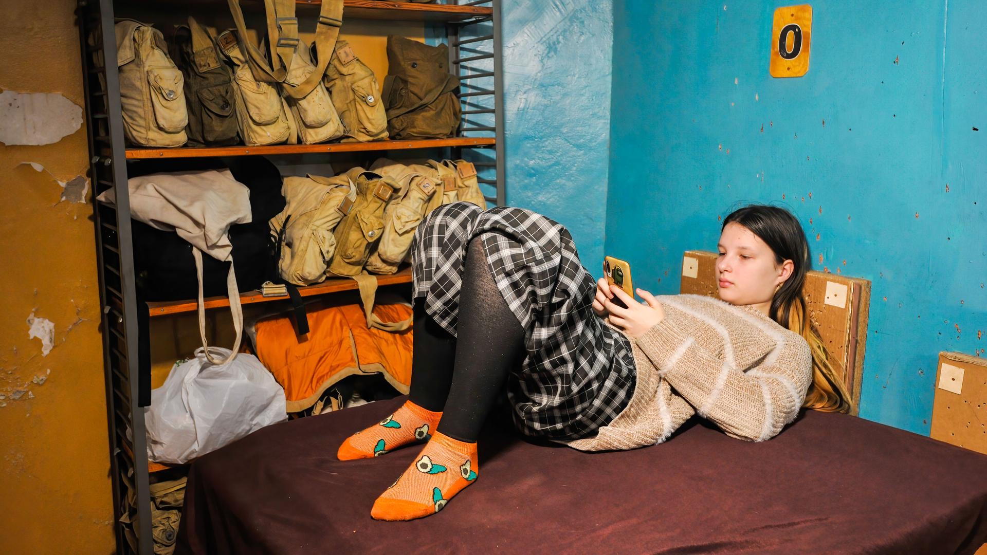 Eine Jugendliche liegt auf einer Liege in einem Luftschutzbunker und schaut auf ihr Smartphone. Eine Jugendliche liegt auf einer Liege in einem Luftschutzbunker und schaut auf ihr Smartphone.
