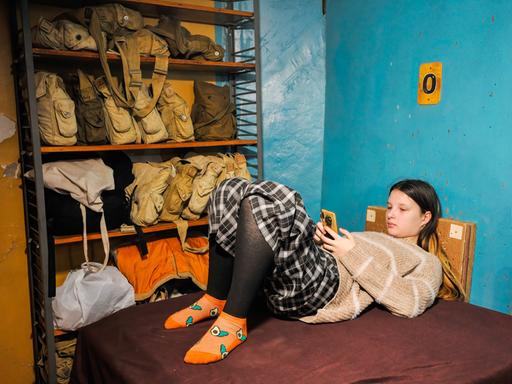 Eine Jugendliche liegt auf einer Liege in einem Luftschutzbunker und schaut auf ihr Smartphone.
