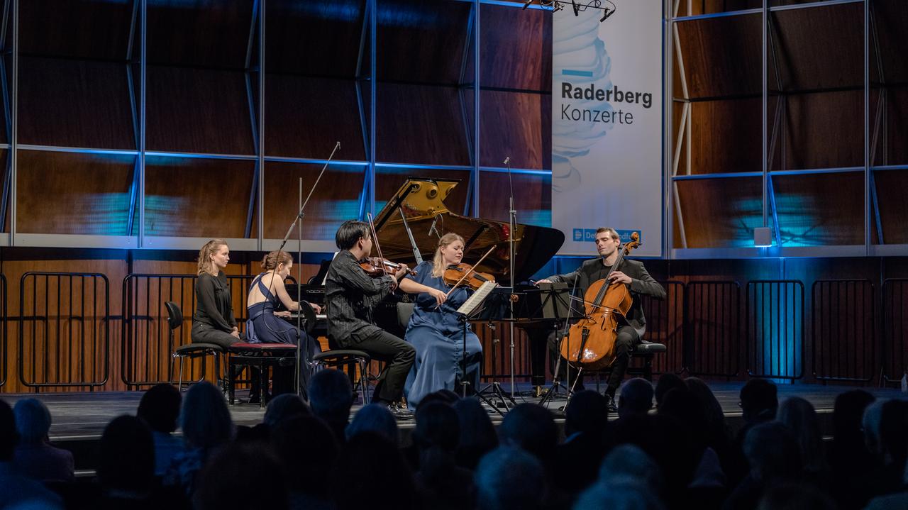 Ein Klavierquartett musiziert auf einem Podium vor Publikum. Der Konzertsaal ist holzgetäfelt und bläulich beleuchtet, die Wand ziert ein Banner mit der Aufschrift "Raderbergkonzerte". Ein Klavierquartett musiziert auf einem Podium vor Publikum. Der Konzertsaal ist holzgetäfelt und bläulich beleuchtet, die Wand ziert ein Banner mit der Aufschrift "Raderbergkonzerte".