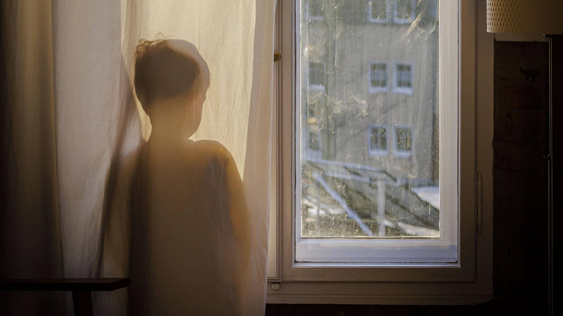 Symbolfoto zum Thema Psychologie bei Kindern. Ein Junge steht am Fenster hinter einem Vorhang und schaut hinaus.