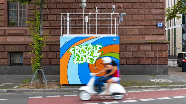 Luftmessstation am Friedrichsring in Mannheim mit Graffiti "Frische Luft" und einem passierenden Roller in der Bewegungsunschärfe. 