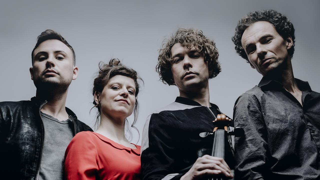 Drei Musiker und eine Musikerin stehen im Halbkreis und blicken zur Kamera, die das Quartett von unten aufnimmt, sodass das Violoncello das Bild dominiert. Drei Musiker und eine Musikerin stehen im Halbkreis und blicken zur Kamera, die das Quartett von unten aufnimmt, sodass das Violoncello das Bild dominiert.