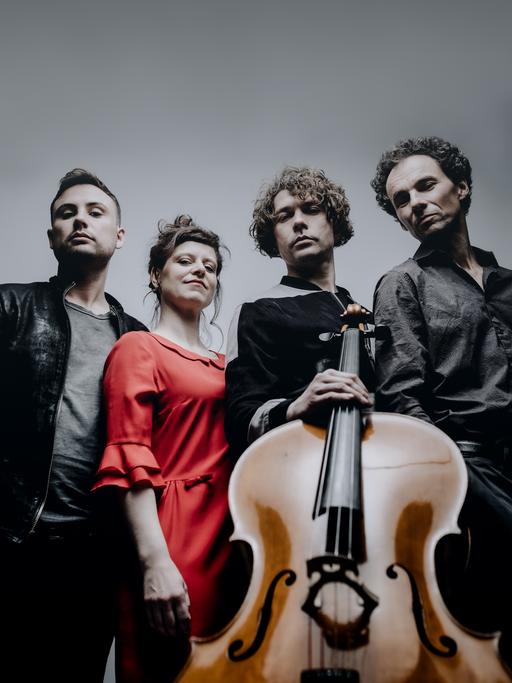 Drei Musiker und eine Musikerin stehen im Halbkreis und blicken zur Kamera, die das Quartett von unten aufnimmt, sodass das Violoncello das Bild dominiert.