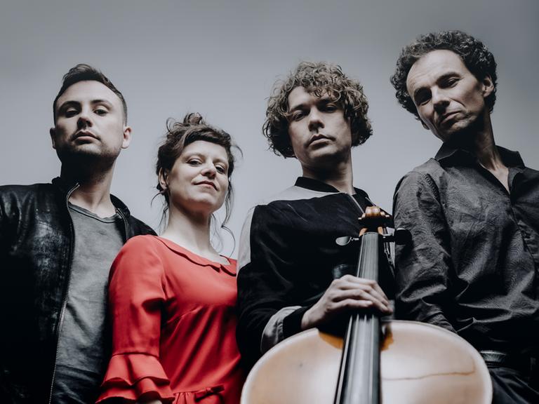Drei Musiker und eine Musikerin stehen im Halbkreis und blicken zur Kamera, die das Quartett von unten aufnimmt, sodass das Violoncello das Bild dominiert.