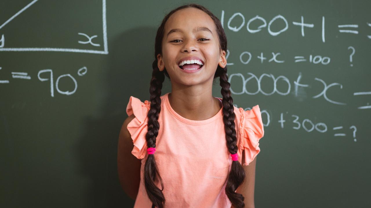 Porträt eines lachenden Schulmädchens. Es steht vor einer Kreidetafel mit Matheaufgaben. 