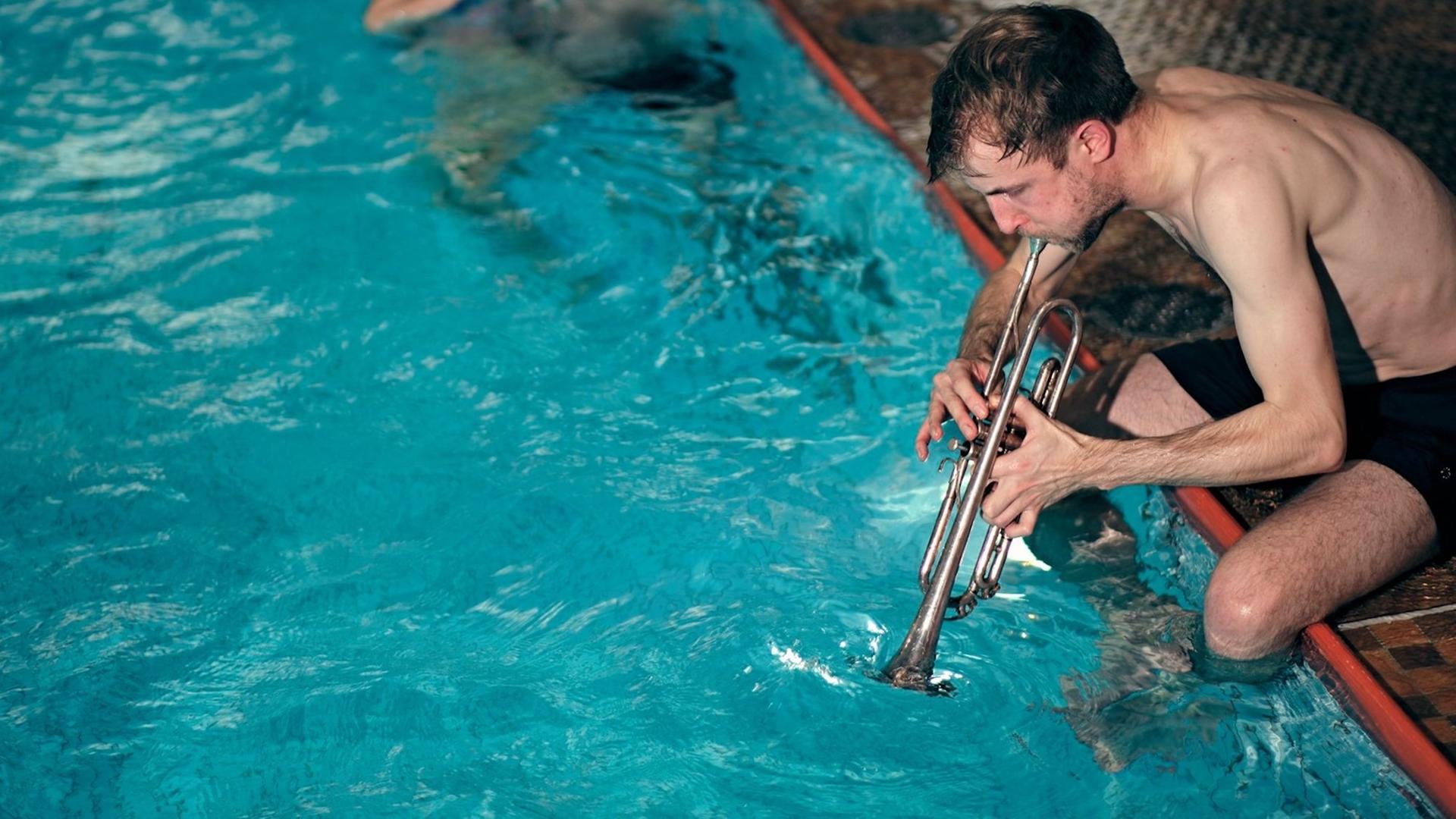 Ein Trompeter in Badehose sitzt am Schwimmbeckenrand und bespielt das Wasser