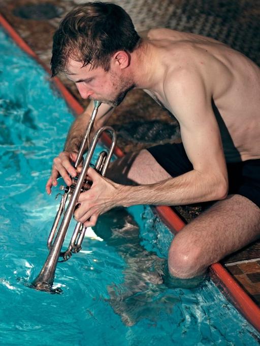 Ein Trompeter in Badehose sitzt am Schwimmbeckenrand und bespielt das Wasser