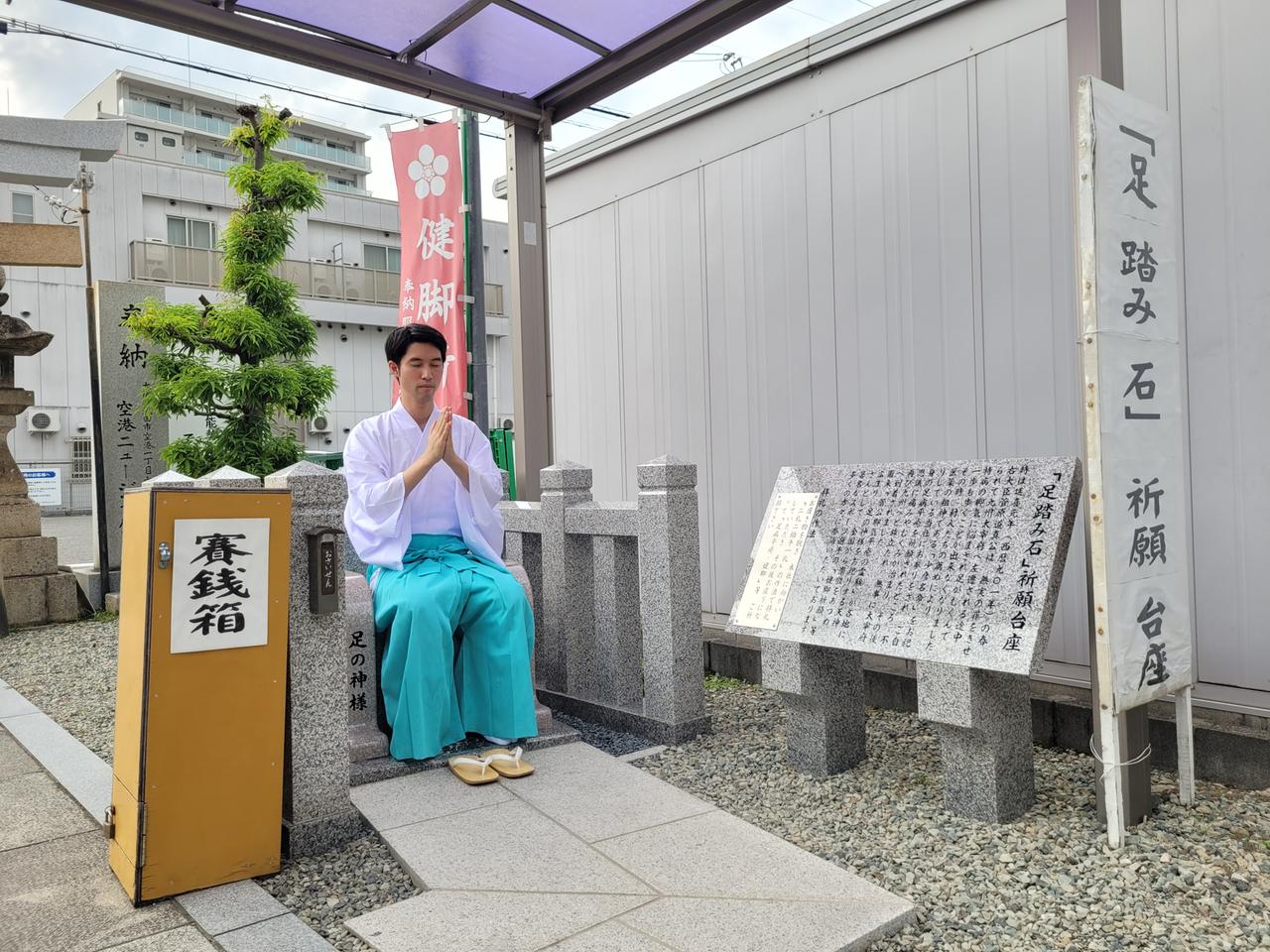 Shinto-Priester Taishi Kato, im weiten weißen Hemd und hellblauem Rock, sitzt auf einem steinernen Gebetsstuhl, er hat die Augen geschlossen, die Hände erhoben und zusammengelegt.
