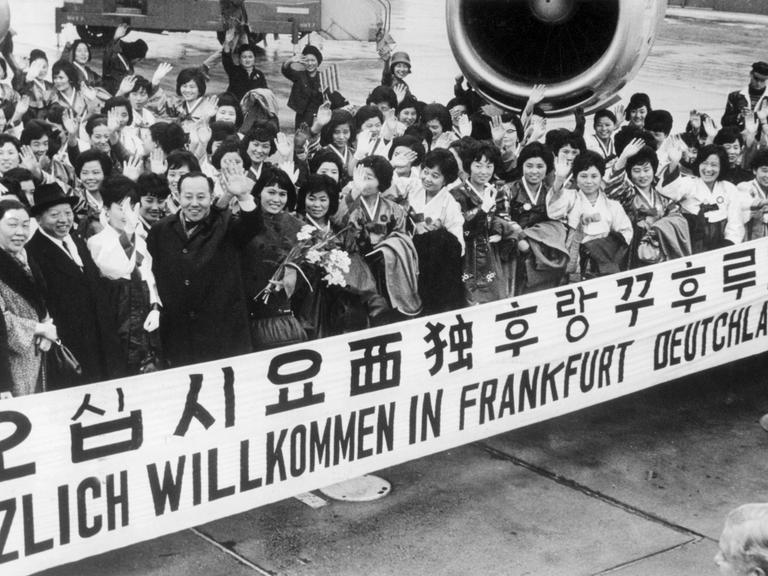 "Herzlich Willkommen in Frankfurt Deutschland" steht auf einem Spruchband, mit dem eine große Gruppe Krankenschwestern aus Südkorea im Jahr 1966 auf dem Rhein-Main-Flughafen begrüßt wird. 