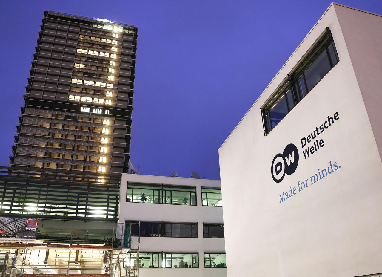 Der Sitz der Deutschen Welle in Bonn: Im Vordergrund rechts ist der Schriftzug des Senders auf einer Hauswand zu sehen, links im Bild steht ein erleuchtetes Hochhaus.