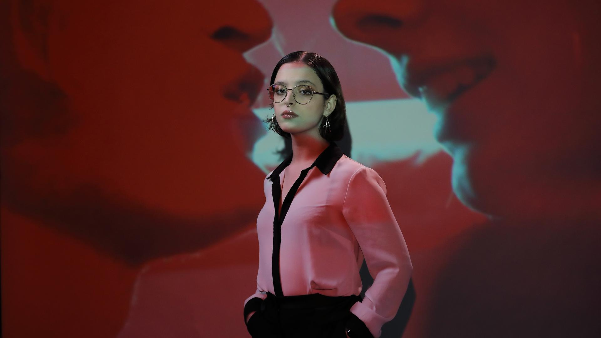 Elizabeth Duval, mit Brille und Kreolen, in einer pinken Bluse und schwarzer Hose, steht vor der Projektion eines Filmausschnitts, in dem zwei Münder einander fast berühren. Elizabeth Duval, mit Brille und Kreolen, in einer pinken Bluse und schwarzer Hose, steht vor der Projektion eines Filmausschnitts, in dem zwei Münder einander fast berühren.