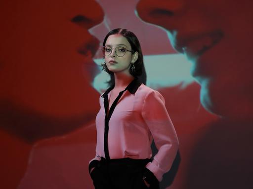 Elizabeth Duval, mit Brille und Kreolen, in einer pinken Bluse und schwarzer Hose, steht vor der Projektion eines Filmausschnitts, in dem zwei Münder einander fast berühren.