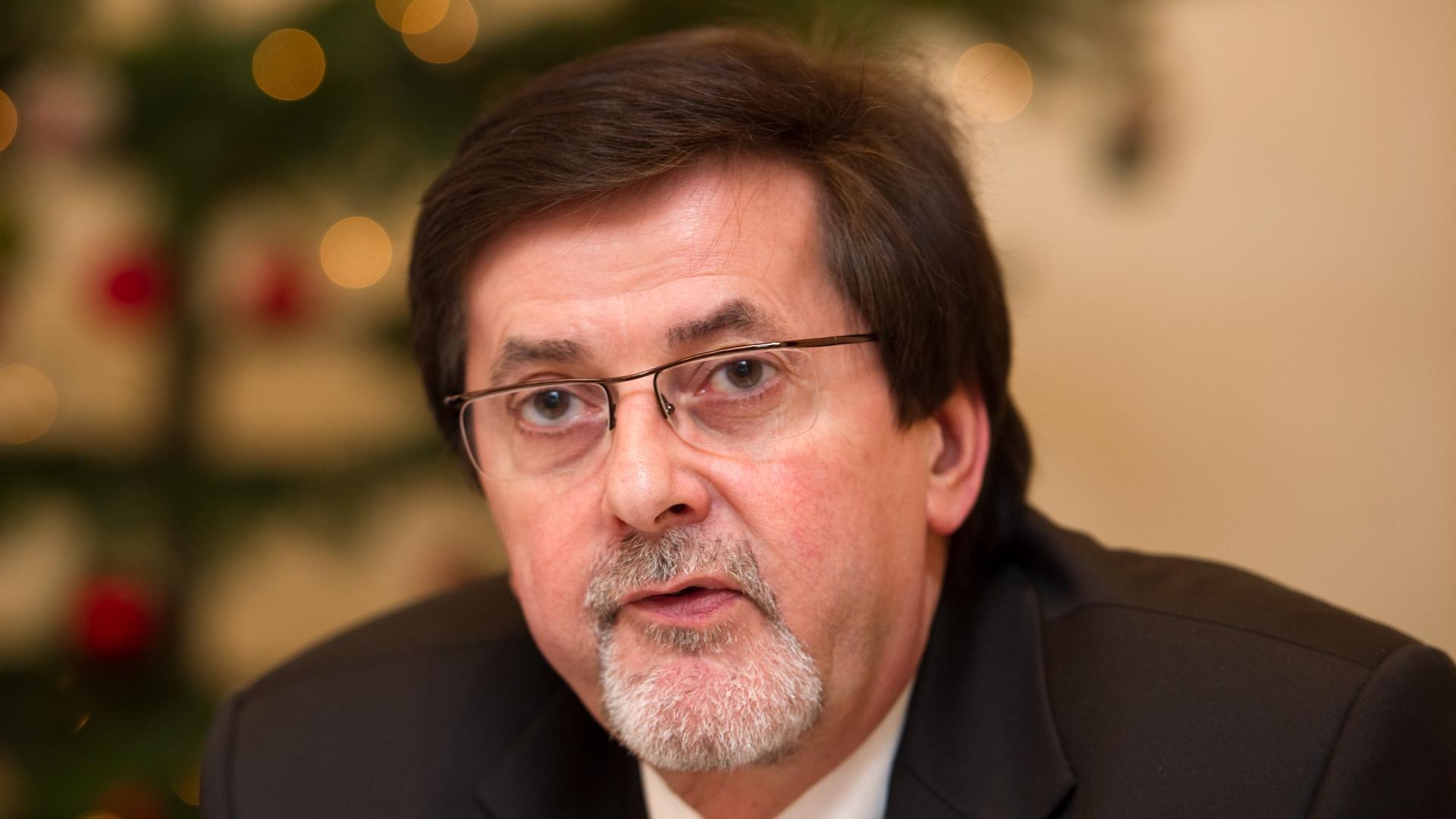 Ein dunkelhaariger Mann mit Brille und Bart schaut ernst, im Hintergrund ist unscharf ein Weihnachtsbaum zu sehen. Ein dunkelhaariger Mann mit Brille und Bart schaut ernst, im Hintergrund ist unscharf ein Weihnachtsbaum zu sehen.