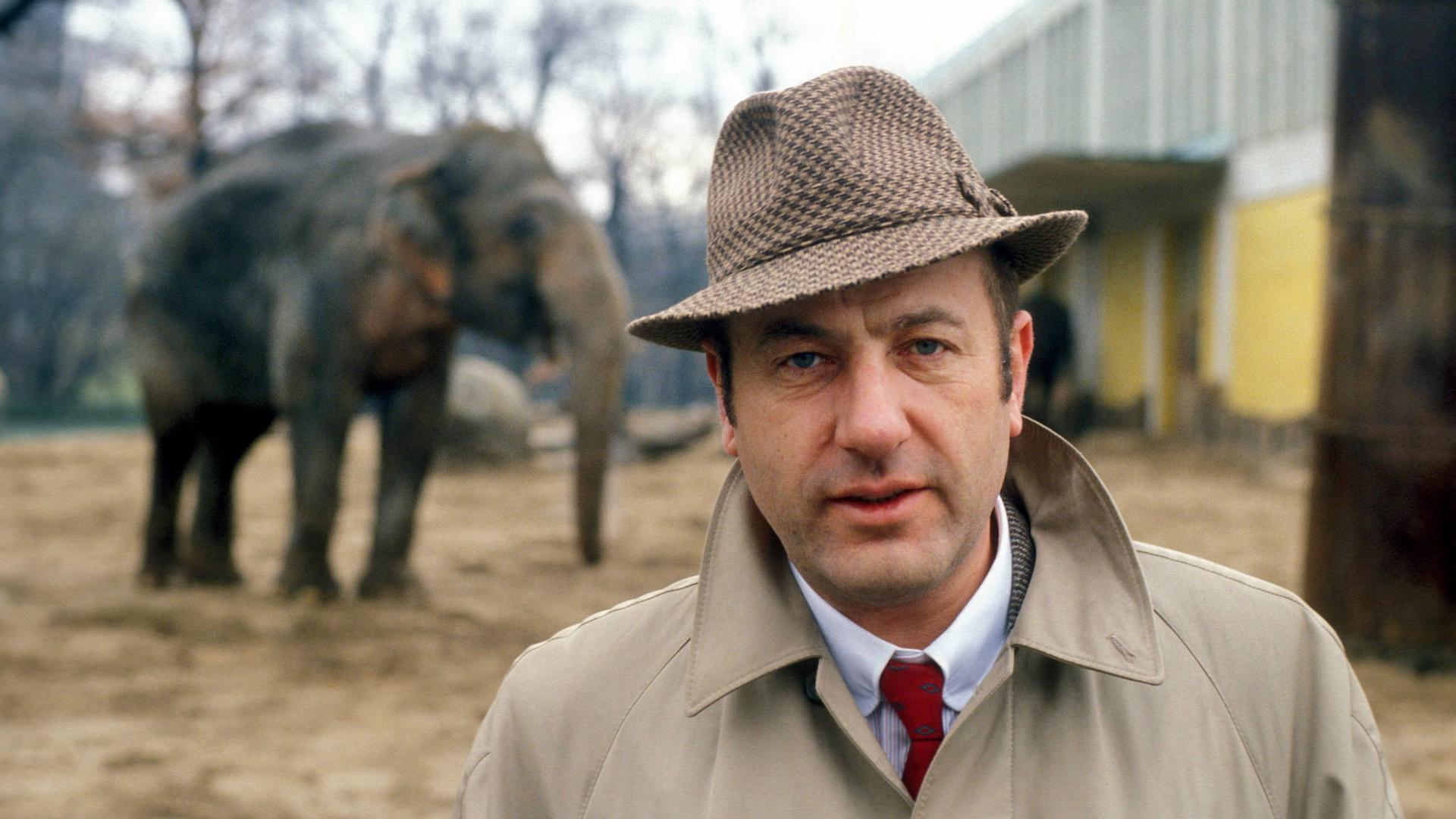 Ein Mann mittleren Alters steht in einem Trenchcoat mit hochgestelltem Kragen und einem karierten Herrenhut, vor dem Außengehege eines Elefanten. Ein Mann mittleren Alters steht in einem Trenchcoat mit hochgestelltem Kragen und einem karierten Herrenhut, vor dem Außengehege eines Elefanten.