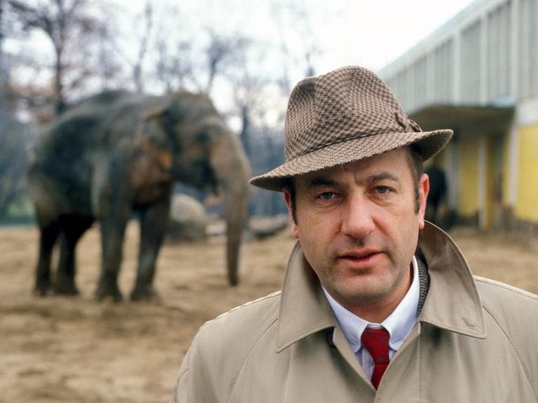 Ein Mann mittleren Alters steht in einem Trenchcoat mit hochgestelltem Kragen und einem karierten Herrenhut, vor dem Außengehege eines Elefanten.
