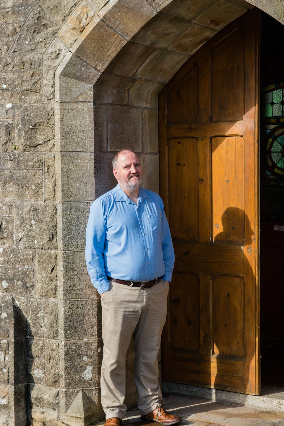 Ein Mann steht, mit Händen in den Hosentaschen, vor einer leicht geöffneten Kirchentür und schaut in die Kamera.