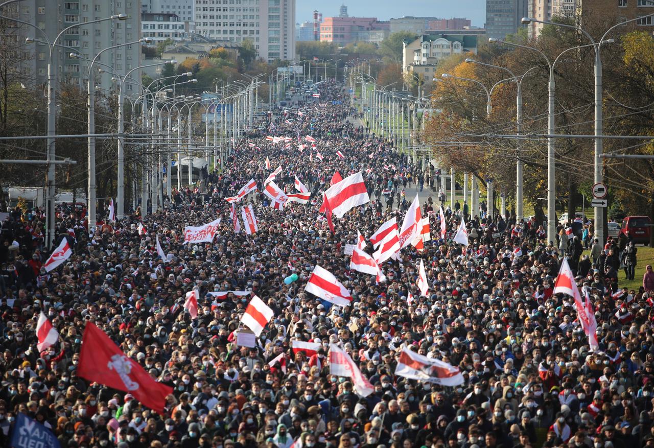 Tausende Menschen mit der belarussischen Flagge in der Hand gehen eine breite Straße in Minsk entlang. 