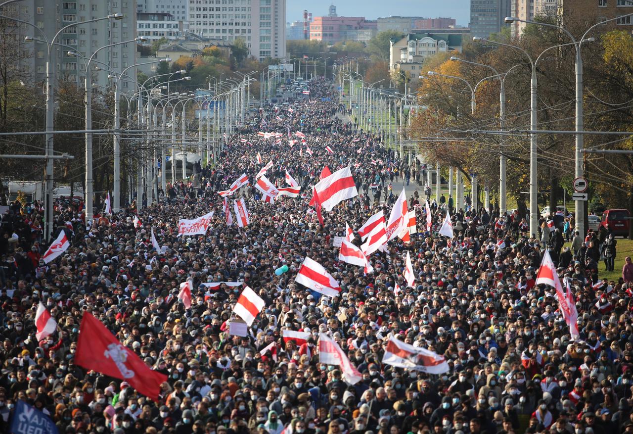 Tausende Menschen mit der belarussischen Flagge in der Hand gehen eine breite Straße in Minsk entlang. Tausende Menschen mit der belarussischen Flagge in der Hand gehen eine breite Straße in Minsk entlang.