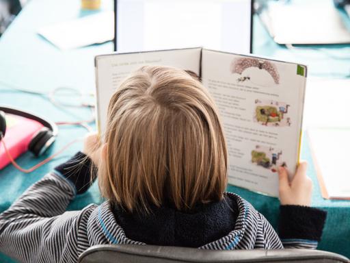 Ein Schulkind hat ein Schulbuch in der Hand, sitzt an einem Tisch und liest. Man sieht es von hinten.