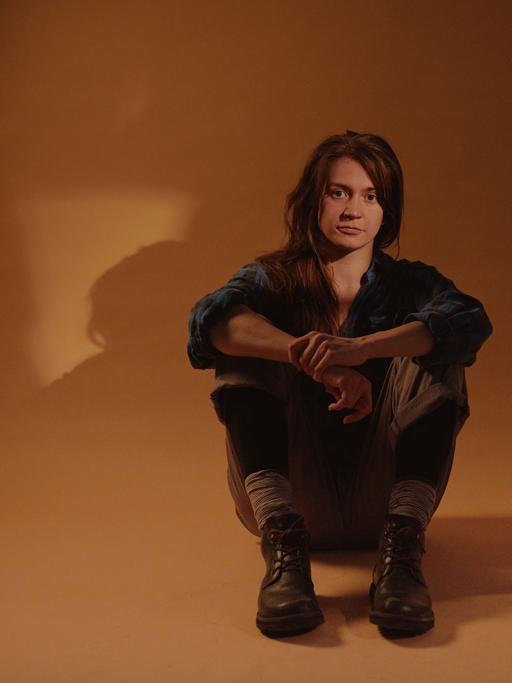 Eine Frau mit langen, braunen Haaren sitzt in Jeans in einem Fotostudio, von Schatten umringt auf gelben Wänden.