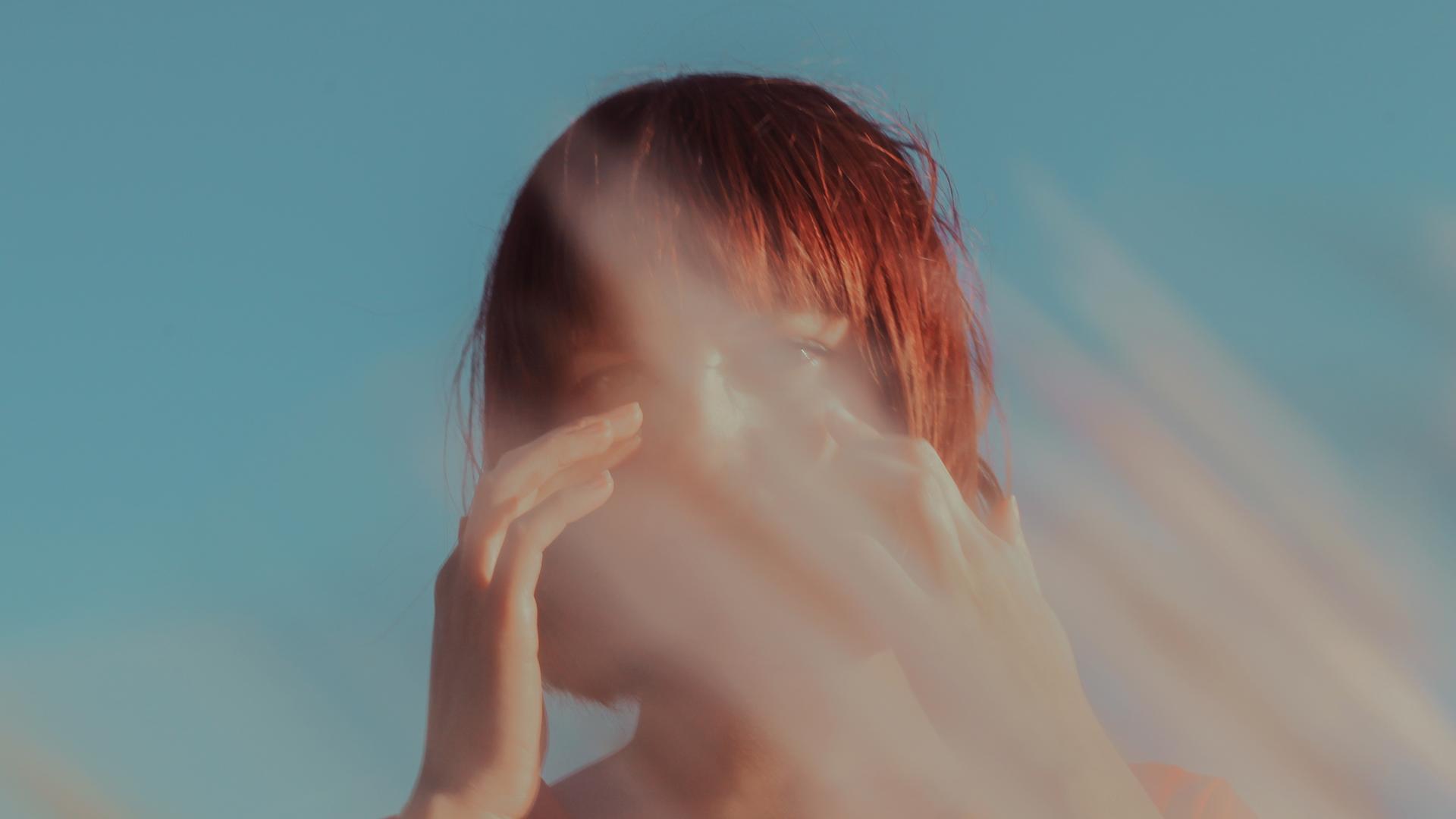Eine junge Frau vor blauem Himmel. Vor ihrem Gesicht unscharfe rötlich-graue Streifen, wie bei Lichtstrahlen, Gräsern oder verwischter Farbe. Eine junge Frau vor blauem Himmel. Vor ihrem Gesicht unscharfe rötlich-graue Streifen, wie bei Lichtstrahlen, Gräsern oder verwischter Farbe.