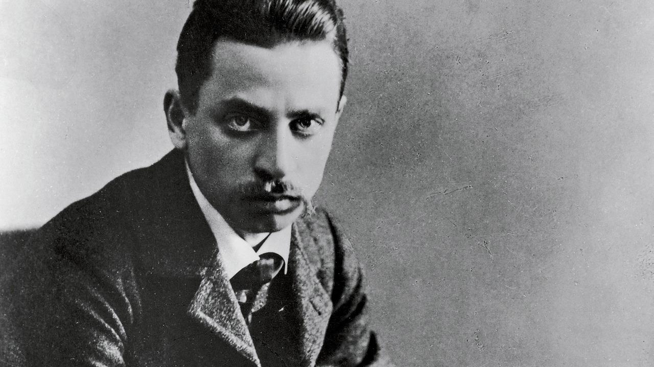 Schwarzweißporträt von Rainer Maria Rilke, der von einem aufgeschlagenen Buch aufblickt. Schwarzweißporträt von Rainer Maria Rilke, der von einem aufgeschlagenen Buch aufblickt.