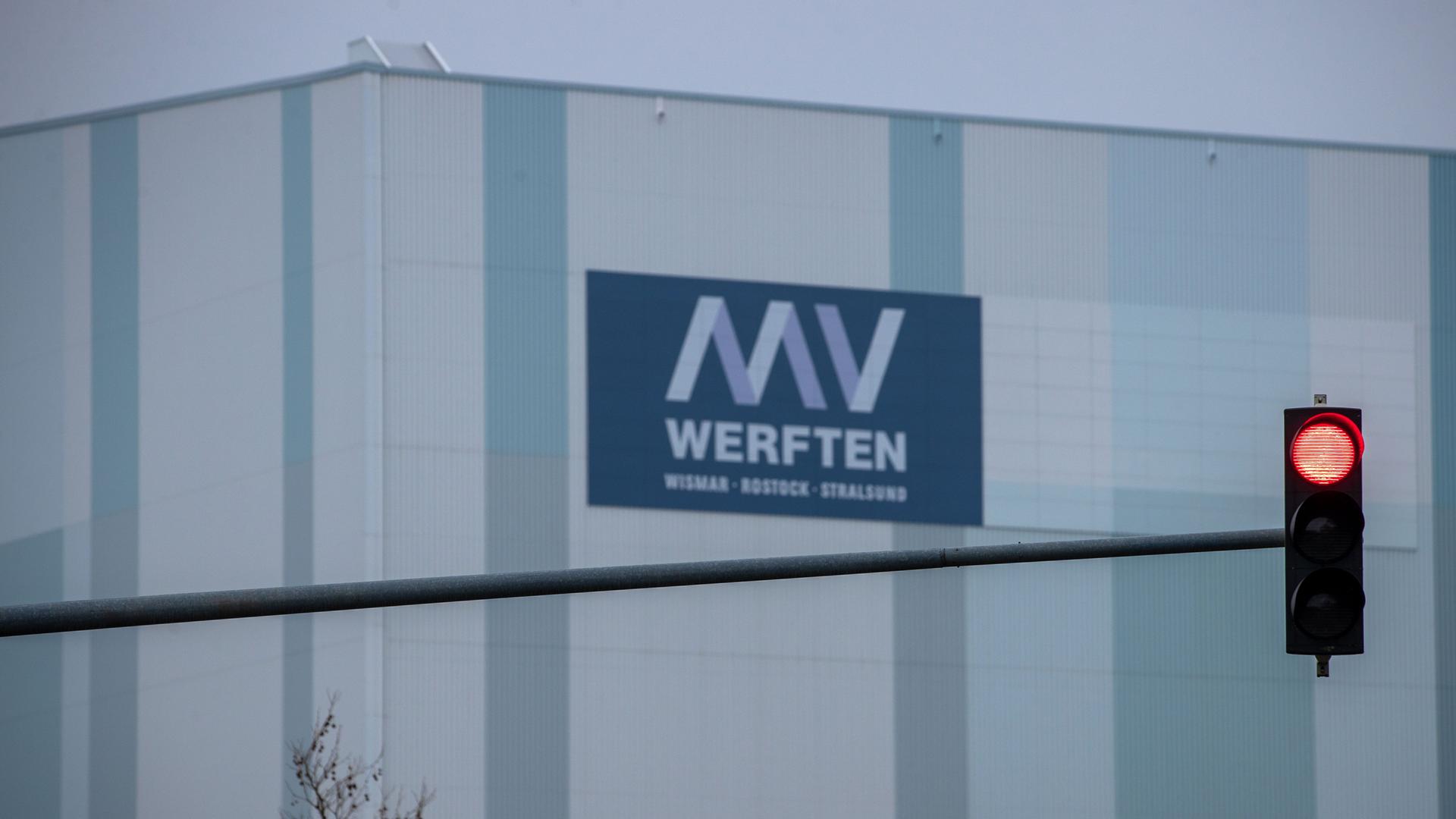 Das ist Gebäude von dem Unternehmen MV Werften. Auf der Wand sieht man das Logo. Davor steht eine rote Ampel. Das ist Gebäude von dem Unternehmen MV Werften. Auf der Wand sieht man das Logo. Davor steht eine rote Ampel.