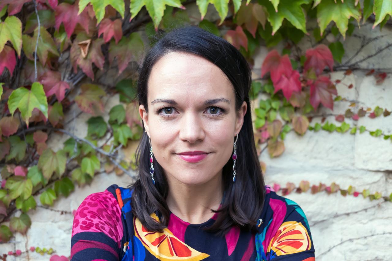 Porträt von Amrei Bahr in einer Bluse mit buntem Muster, mit grünem und rotem Weinlaub im Hintergrund