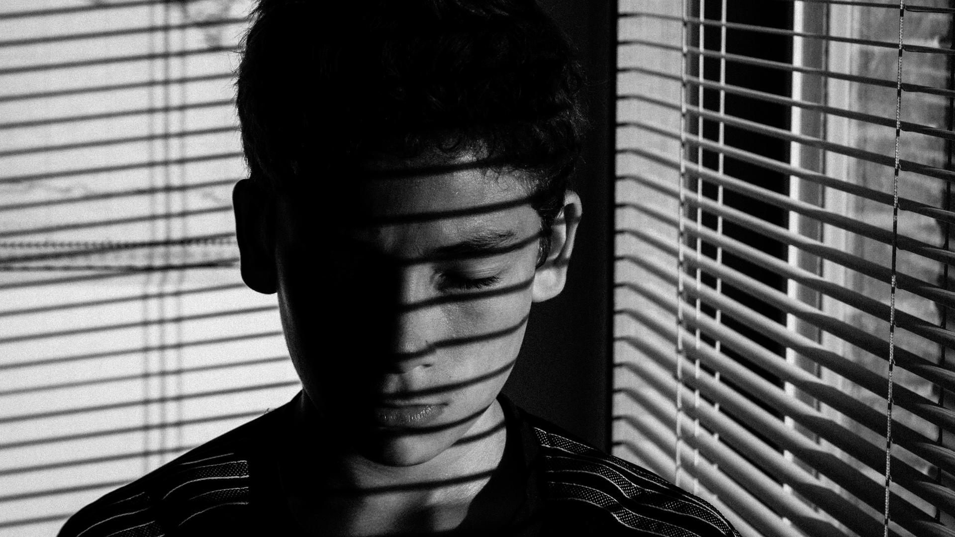 "Fremd" ist eine Geschichte vom Aufwachsen in Widersprüchen. Zu sehen: Ein Junge in einem Raum mit halb geschlossenen Jalousien. "Fremd" ist eine Geschichte vom Aufwachsen in Widersprüchen. Zu sehen: Ein Junge in einem Raum mit halb geschlossenen Jalousien.