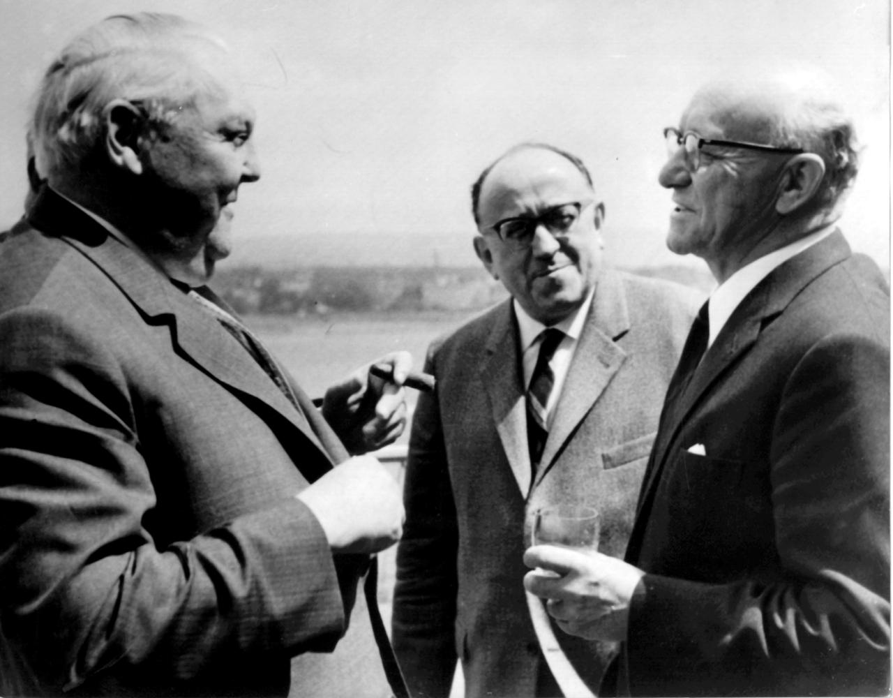 Ludwig Erhard, Dr. Alfred Müller Armack und Dr. Nicholaas Diederichs stehen sich unterhaltend nebeneinander. Erhard hat eine Zigarre in der Hand, Diedrichs ein Glas Wein.