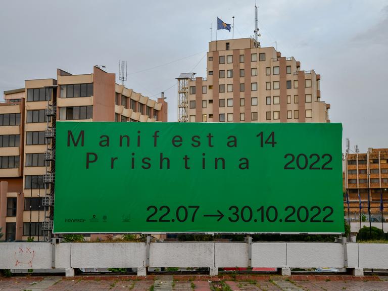 Ein großes günes Plakat mit der Aufschrift "Manifesta 14 / Prishtina 2022" hängt vor einem Wohnblock.
