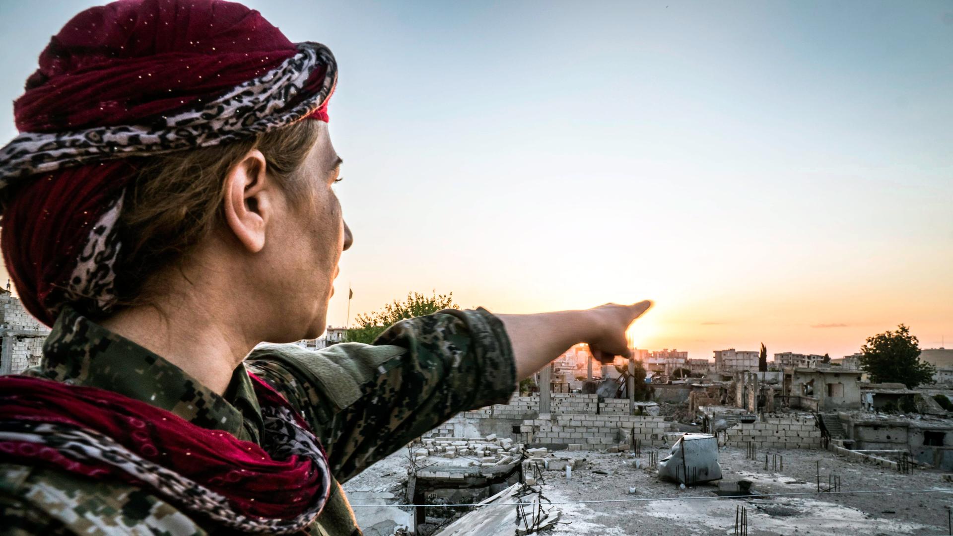 Eine kämpfende Frau in Rojava, Syrien mit Turban zeigt auf Häuser in der Ferne.