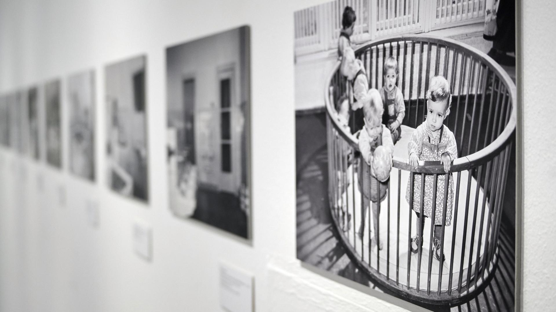 Fotos von kleinen Kindern in einem Gitterbett in der Ausstellung "Wochenkrippen in der DDR" in der Kunsthalle Rostock. Die Ausstellung zeigt rund 20 Kunstwerke zum Thema, ergänzt durch Fotografien und Objekte aus ehemaligen Wochenkrippen sowie Texttafeln und Filme.