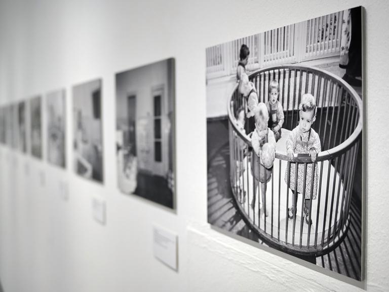 Fotos von kleinen Kindern in einem Gitterbett in der Ausstellung "Wochenkrippen in der DDR" in der Kunsthalle Rostock. Die Ausstellung zeigt rund 20 Kunstwerke zum Thema, ergänzt durch Fotografien und Objekte aus ehemaligen Wochenkrippen sowie Texttafeln und Filme.