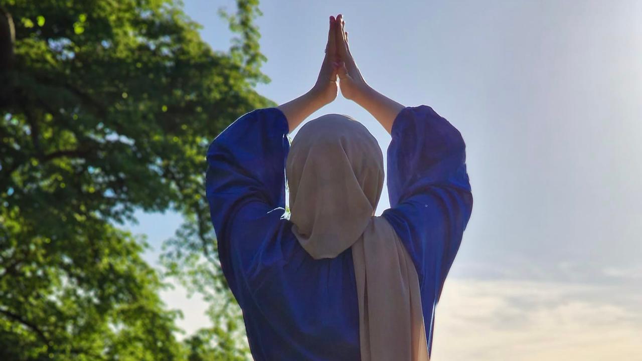Eine Frau mit einem Kopftuch von hinten. Sie steht auf einer Wiese, winkelt ein Bein an und stützt den Fuß gegen das andere Bein. Sie hat die Hände gefaltet über dem Kopf. Eine Frau mit einem Kopftuch von hinten. Sie steht auf einer Wiese, winkelt ein Bein an und stützt den Fuß gegen das andere Bein. Sie hat die Hände gefaltet über dem Kopf.