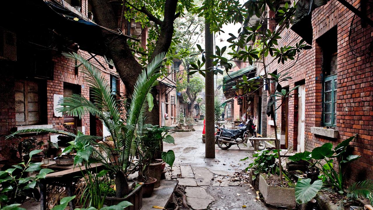 Blick in die Siedlung Tongyuanju in Chongqing, auf beiden Seiten eines schmalen Wegs rote Ziegelsteinhäuser, davor ein Motorrad und Pflanzen.