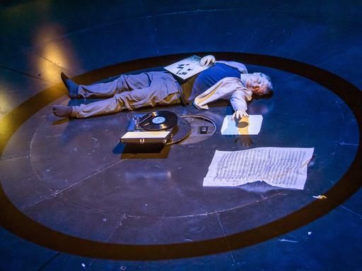 Ein Mann liegt im Inneren eines aufgemalten Kreises auf der Bühne. Vor ihm ein Plattenspieler. Auf seinem Bauch eine Schallplatte.
