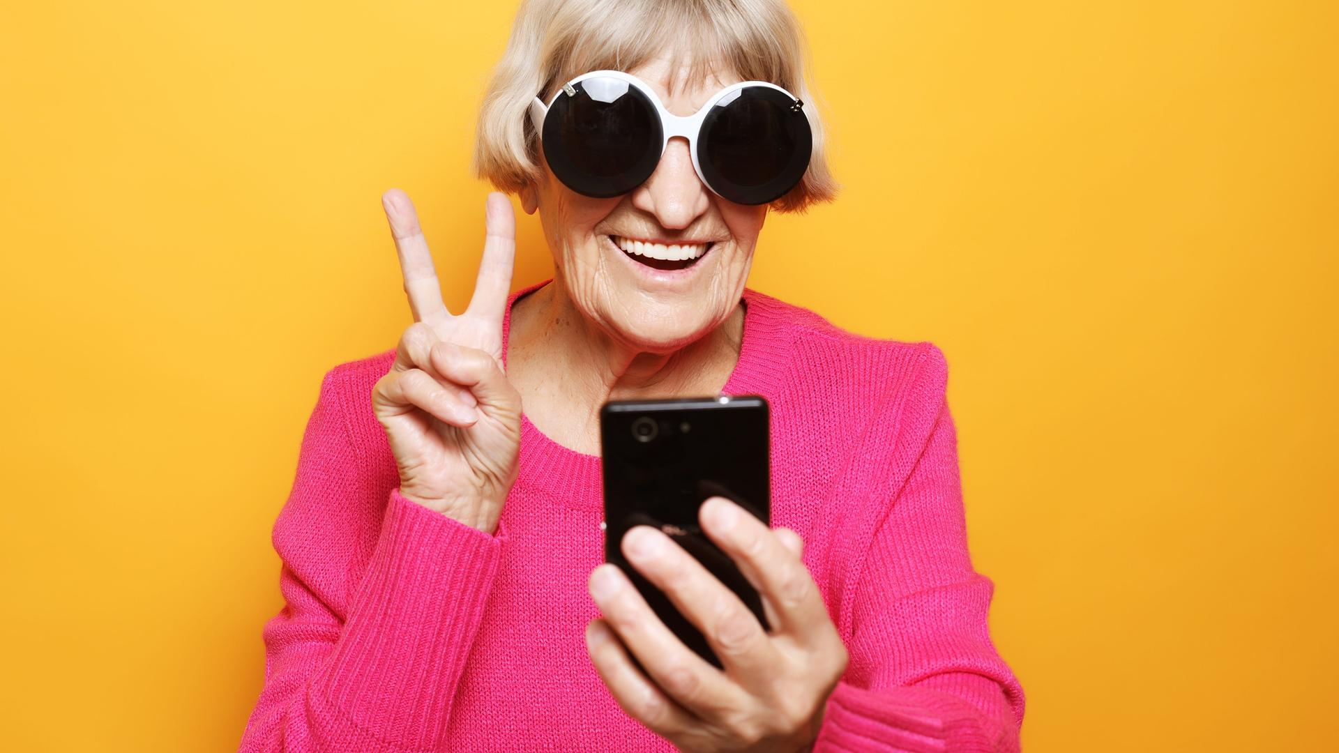 Eine ältere Frau mit einer übergroßen Brille macht ein Victory-Zeichen mit den Fingern und lacht in die Selfiekamera ihres Smartphones.