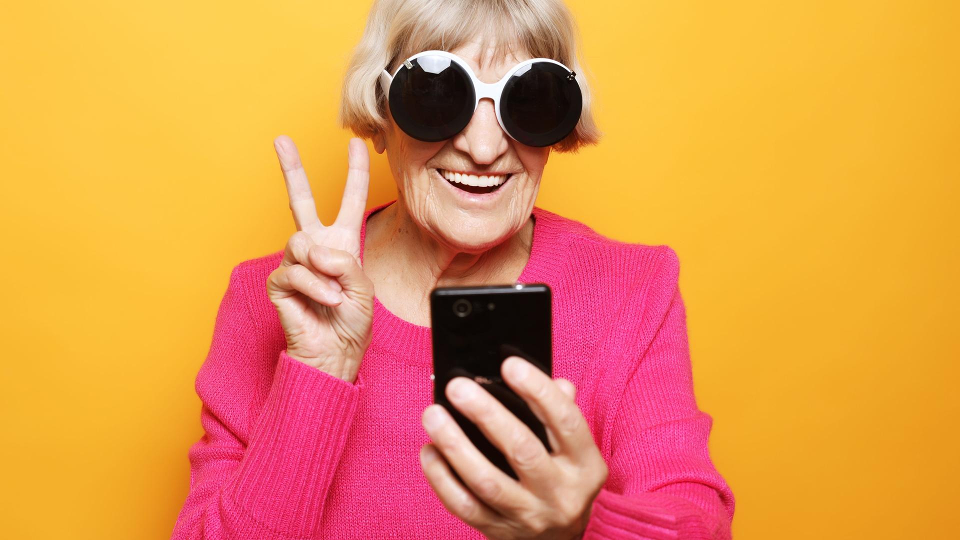 Eine ältere Frau mit einer übergroßen Brille macht ein Victory-Zeichen mit den Fingern und lacht in die Selfiekamera ihres Smartphones. Eine ältere Frau mit einer übergroßen Brille macht ein Victory-Zeichen mit den Fingern und lacht in die Selfiekamera ihres Smartphones.