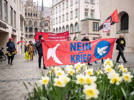 Einige Demonstranten tragen ein rotes Banner mit der Aufschrift "Nein zum Krieg" beim Ostermarsch durch die Münchner Innenstadt. Im Vordergrund blühen Osterglocken. 