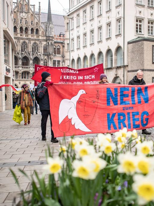 Einige Demonstranten tragen ein rotes Banner mit der Aufschrift "Nein zum Krieg" beim Ostermarsch durch die Münchner Innenstadt. Im Vordergrund blühen Osterglocken. 