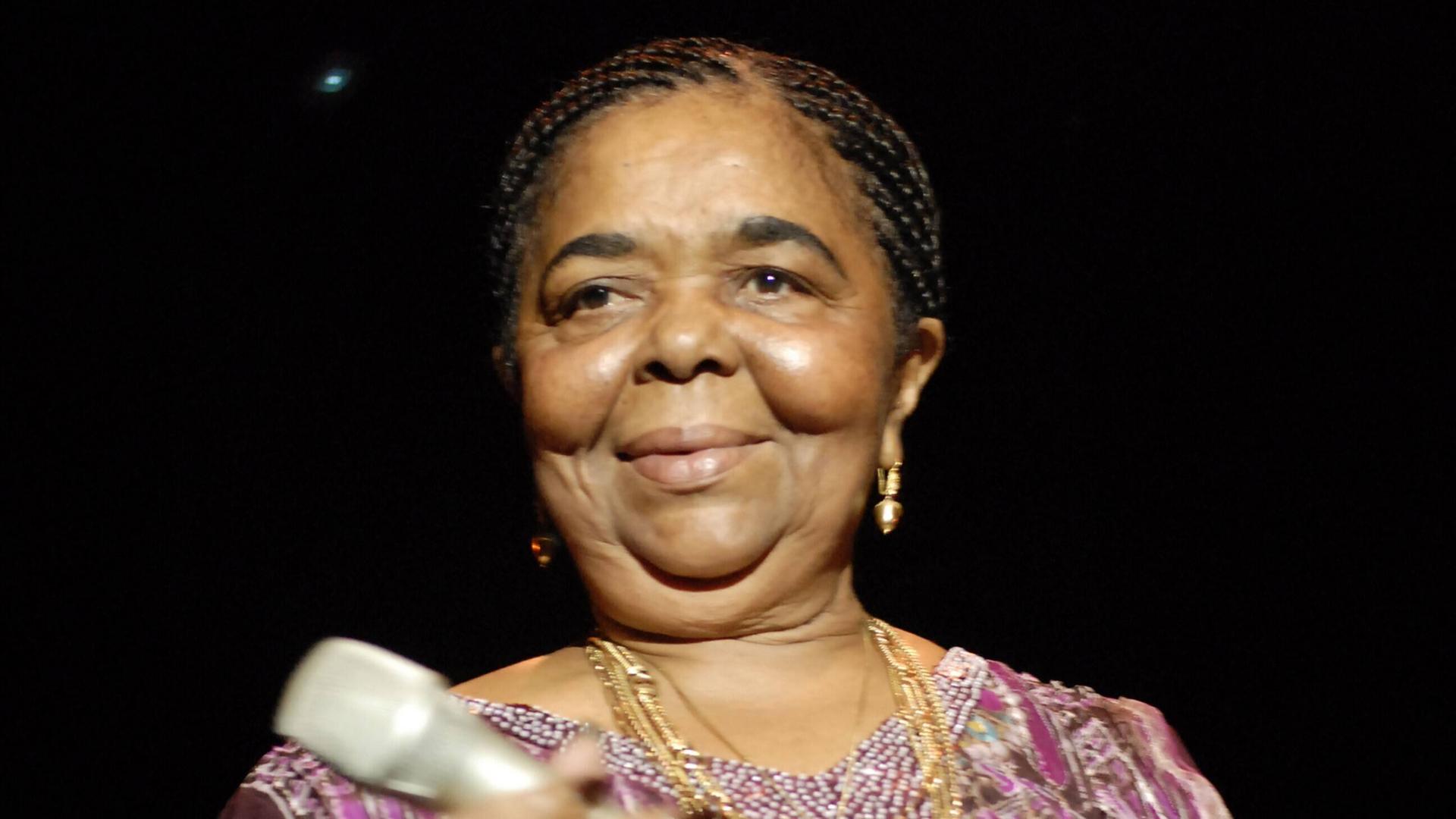 2006 steht Cesária Évora in einer lilafarbenen Bluse mit Mikrophon in den Händen auf einer Bühne und lächelt. 2006 steht Cesária Évora in einer lilafarbenen Bluse mit Mikrophon in den Händen auf einer Bühne und lächelt.