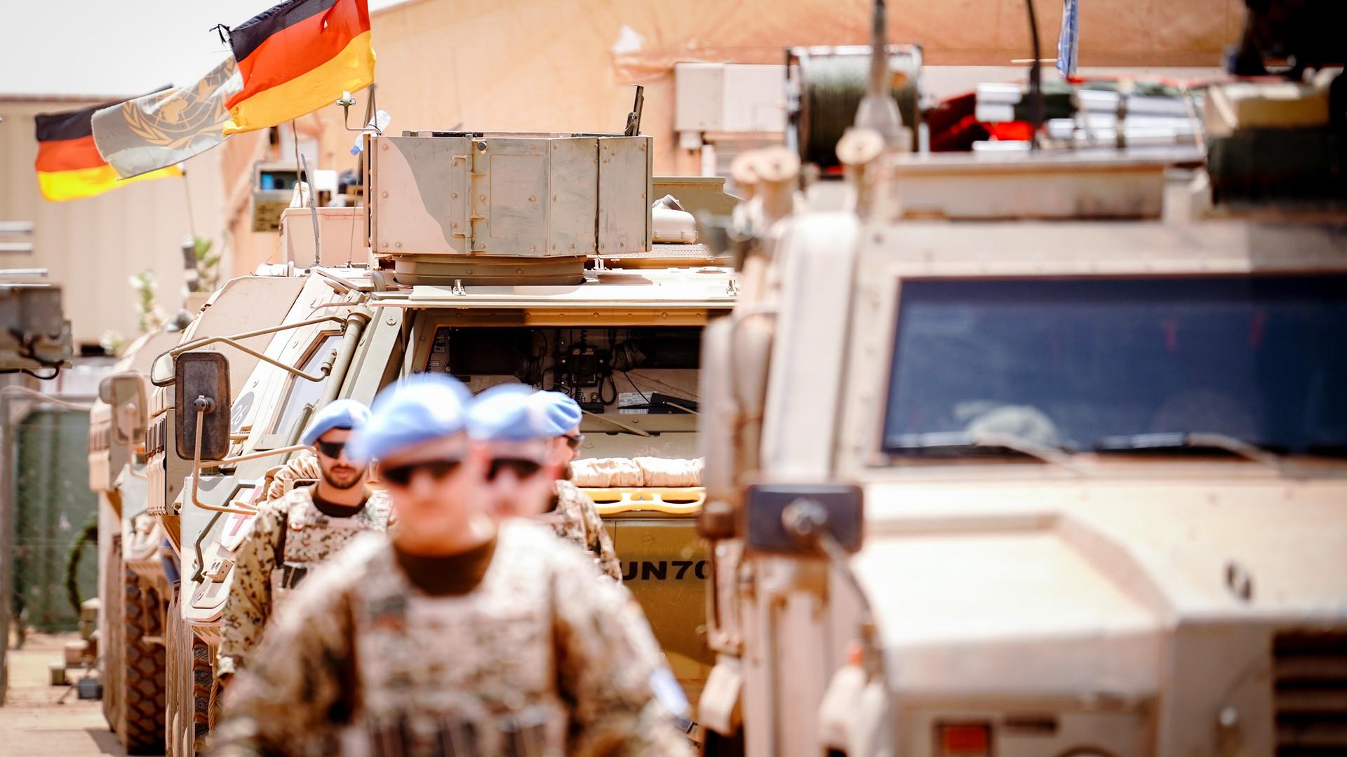 Die Deutschlandfahne und die Flagge der Vereinten Nationen wehen im Camp Castor in Gao, im Vordergrund sind Soldaten und gepanzerte Fahrzeuge zu sehen.
