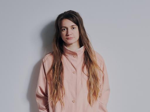 Katharina Kollmann steht in einer rosafarbenen Jacke und offenem, braunen Haar an eine Wand gelehnt und schaut in die Kamera.