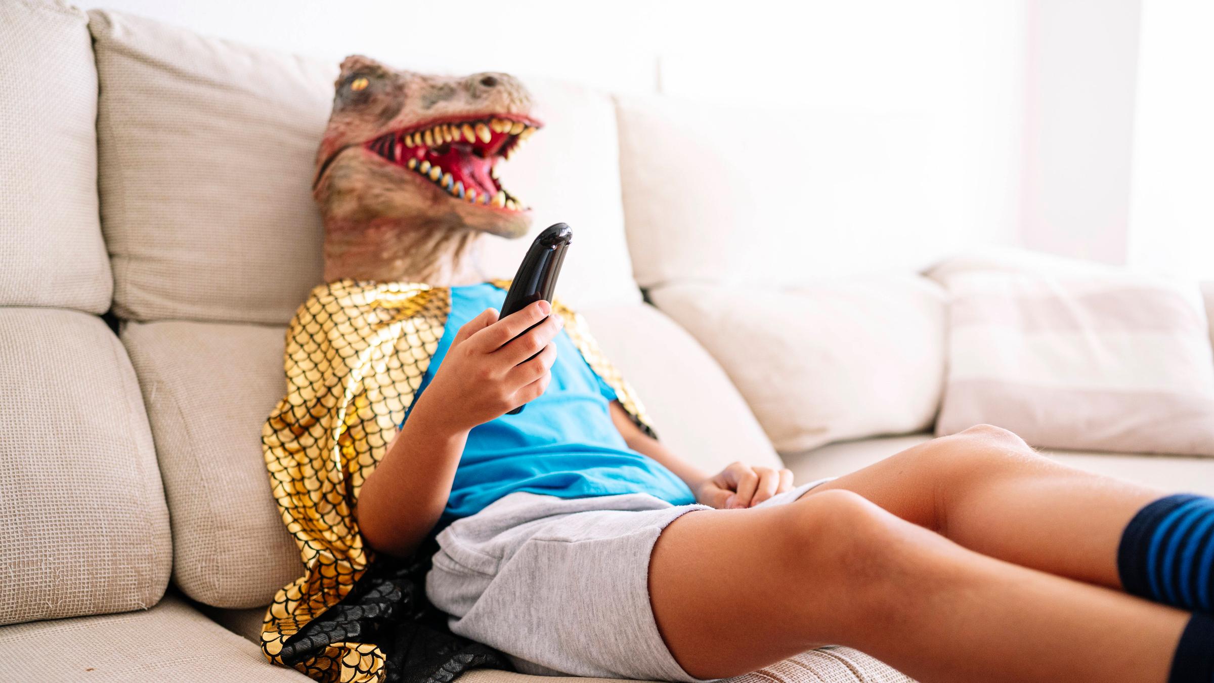 Ein Kind trägt eine Dinosauriermaske und sitzt mit der TV Fernbedienung in der Hand auf einem Sofa.