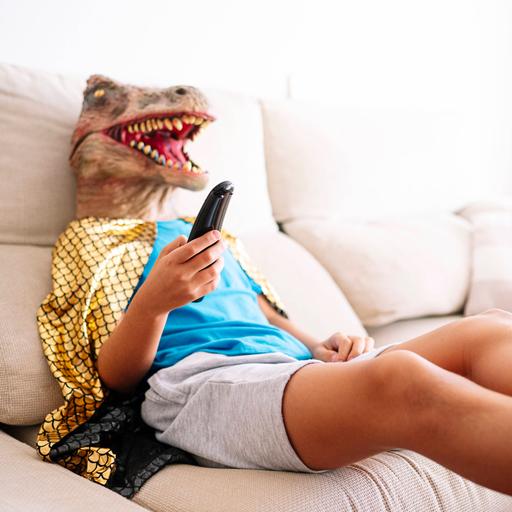 Ein Kind trägt eine Dinosauriermaske und sitzt mit der TV Fernbedienung in der Hand auf einem Sofa.