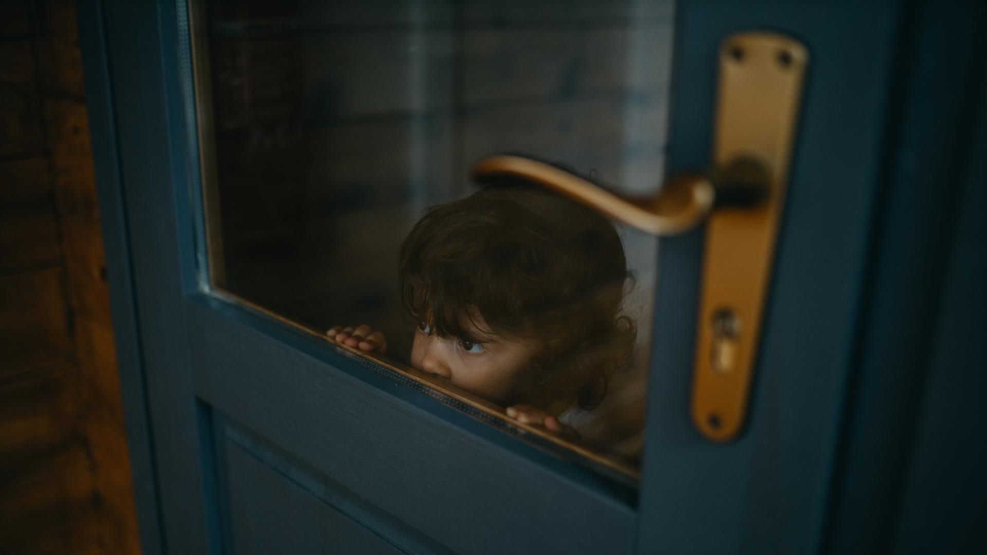 Ein Kind schaut durch die Glasscheibe einer blauen Holztür. Es reicht mit der Nase gerade bis zu der unteren Kante der Scheibe. Es sieht aus, als würde es sich verstecken.